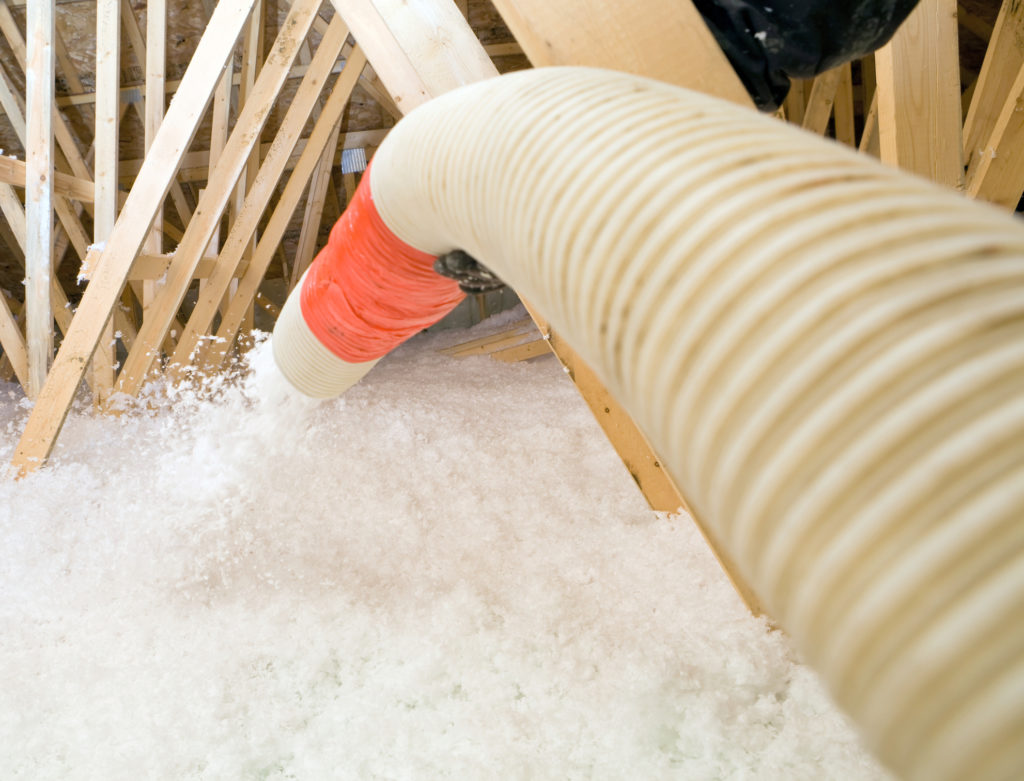 A worker is spraying blown fiberglass insulation between attic trusses at a residential construction site.