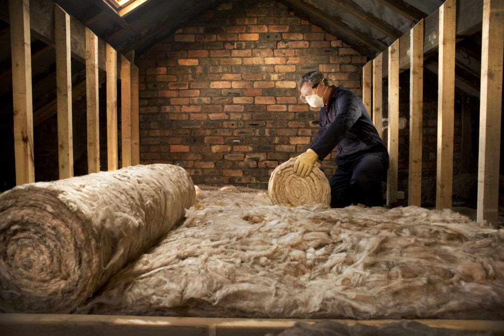 A man putting insulation down in his roof with gloves, eye goggles and a mask on.